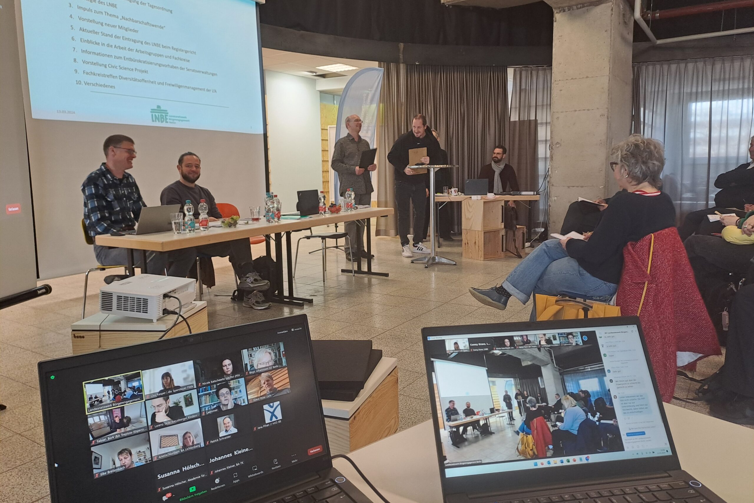 Zwei Laptops im Vordergrund zeigen die digitalen Teilnehmer der Mitgliederversammlung. Im Hintergrund ist eine Power Point Präsentation zu sehen sowie der Sprecher:innenrat.