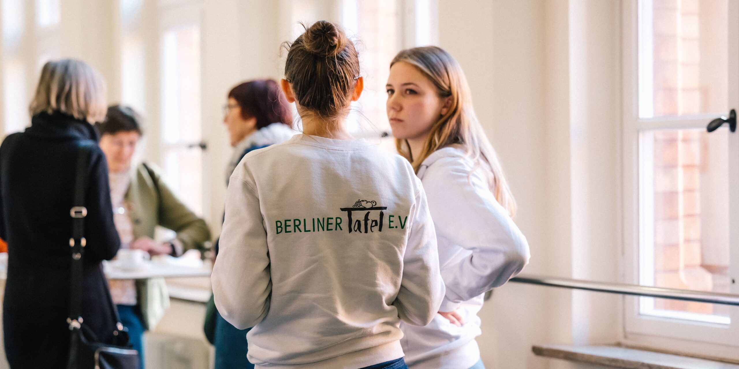 Zwei junge Frauen unterhalten sich. Die eine steht mit dem Rücken zum Bild, auf dem das Logo der Berliner Tafel abgedruckt ist.