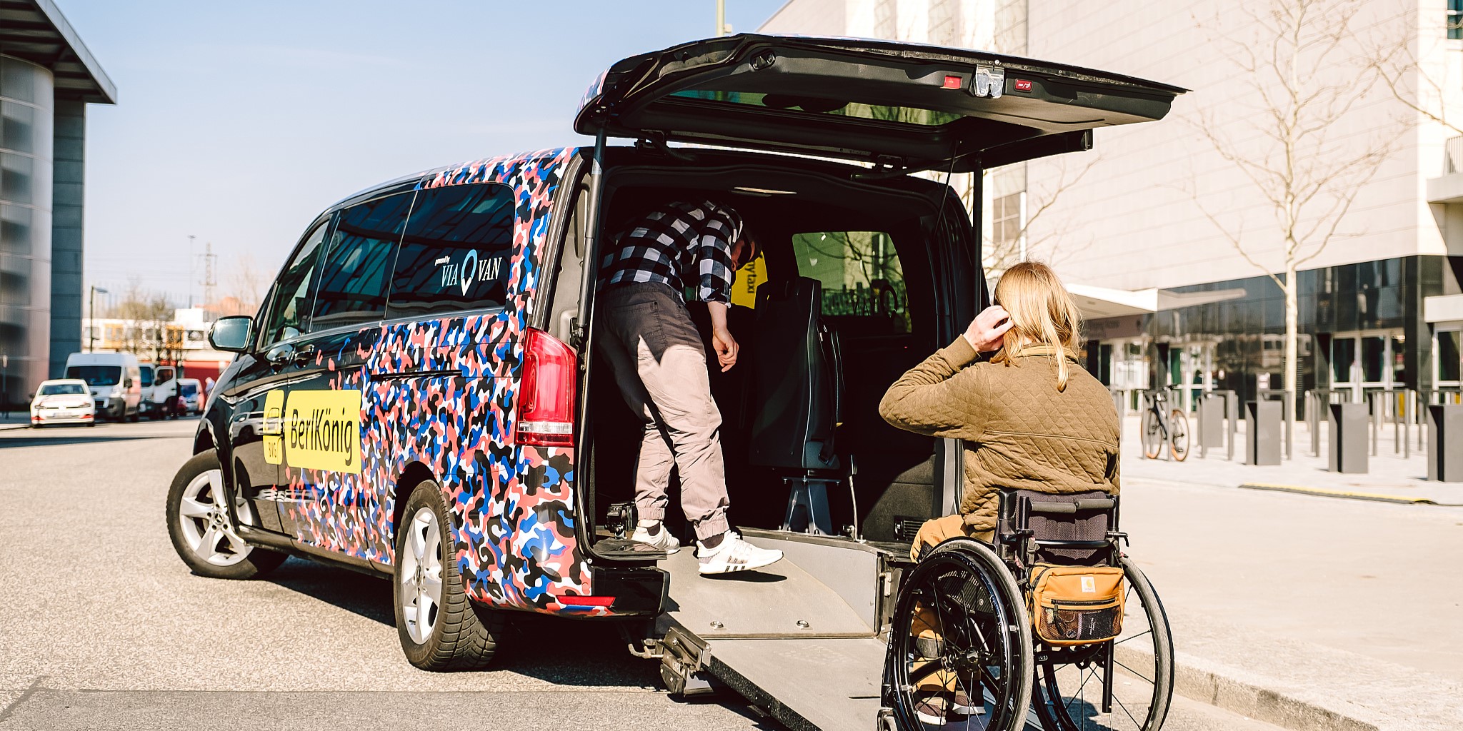 Im Bild ist ein rollstuhlgerechter Van des Tochterunternehmens der BVG namens 'Berlkönig' zu sehen. Vor der Rampe wartet eine Person im Rollstuhl, während eine weitere Person den Einstieg vorbereitet.