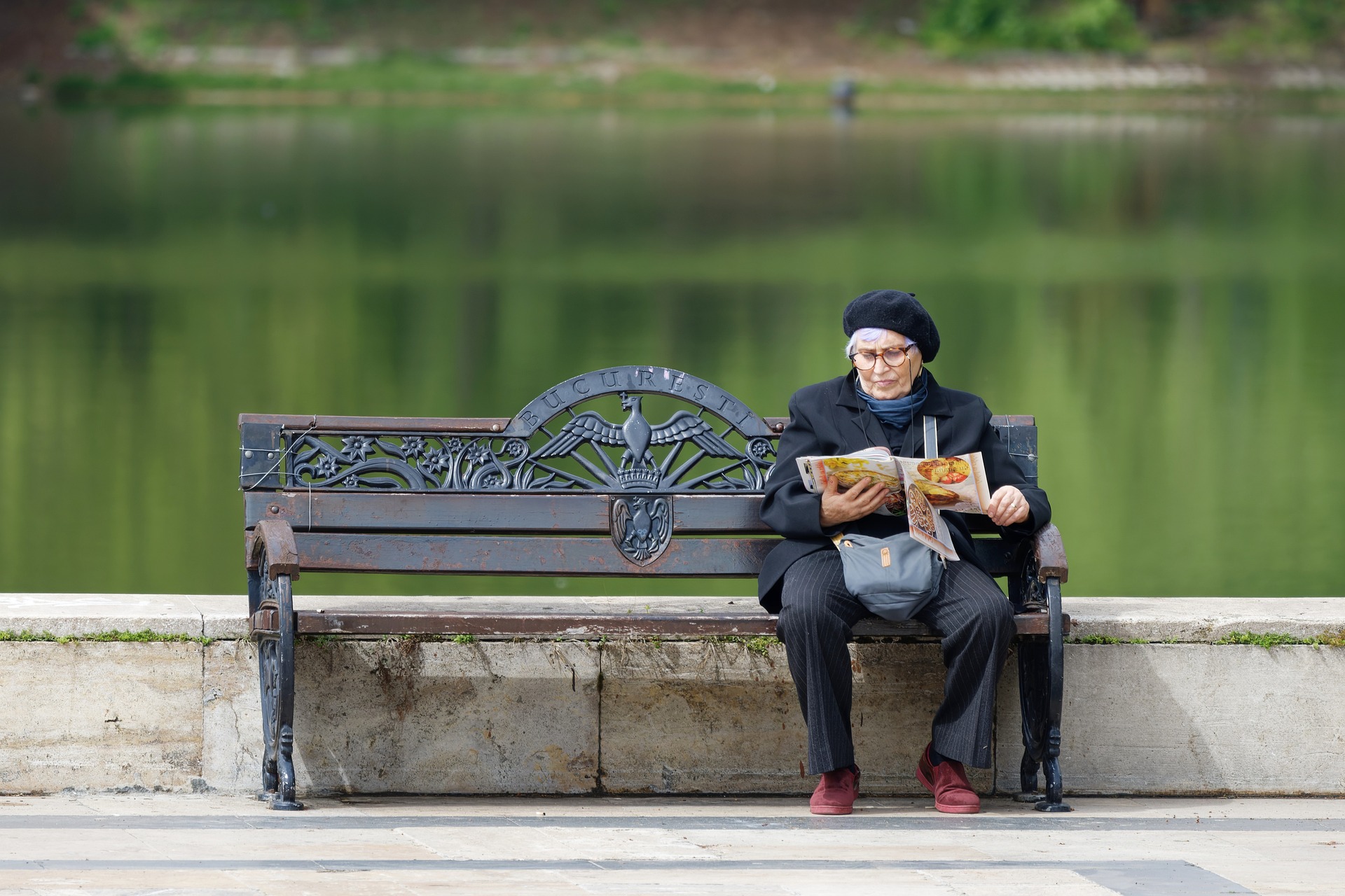 Eine ältere Frau sitzt auf einer Parkbank und blättert durch eine Zeitung. Im Hintergrund ist ein See zu sehen.
