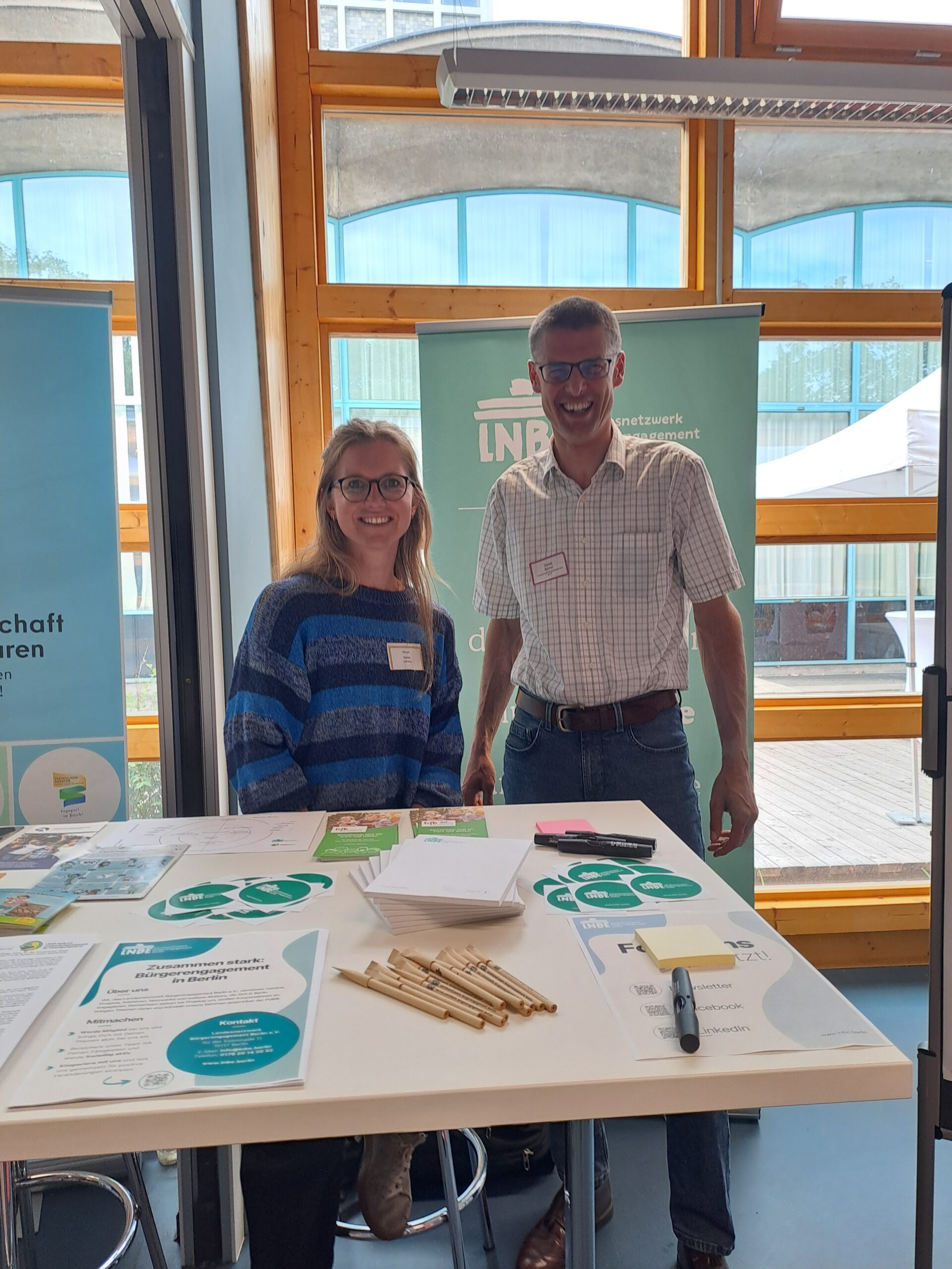 Eine Frau und ein Mann posieren hinter dem Infostand des Landesnetzwerks Bürgerengagement Berlin. im Hintergrund ist ein Roll-Up zu sehen.