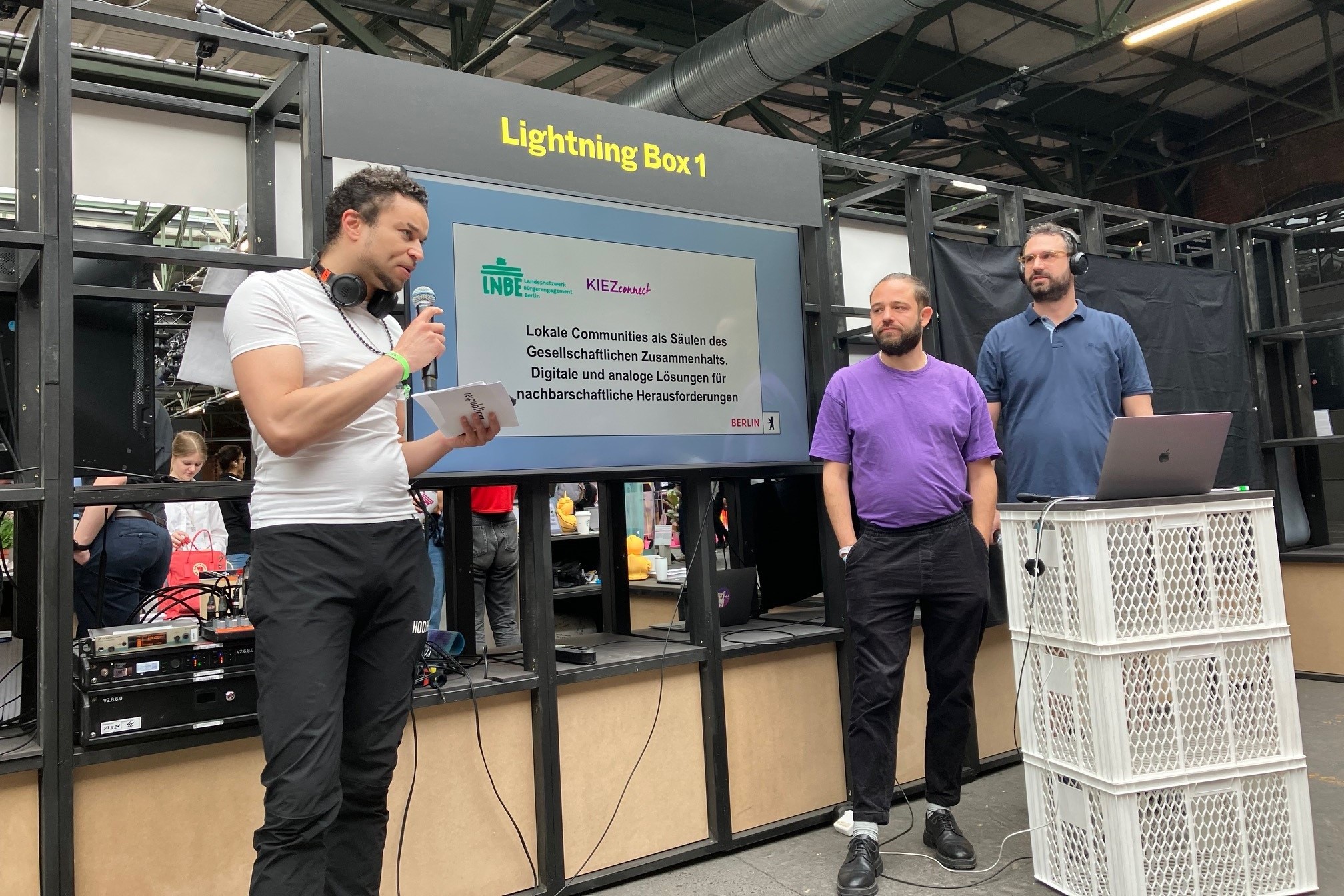 Ein Präsentationsstand auf der Konferenz re:publica. Ein Moderator in einem weißen T-Shirt kündigt die nächste Präsentation an, während zwei Männer in blauen und lila T-Shirts bereitstehen. Im Hintergrund ist die Präsentation auf einem Bildschirm zu sehen.