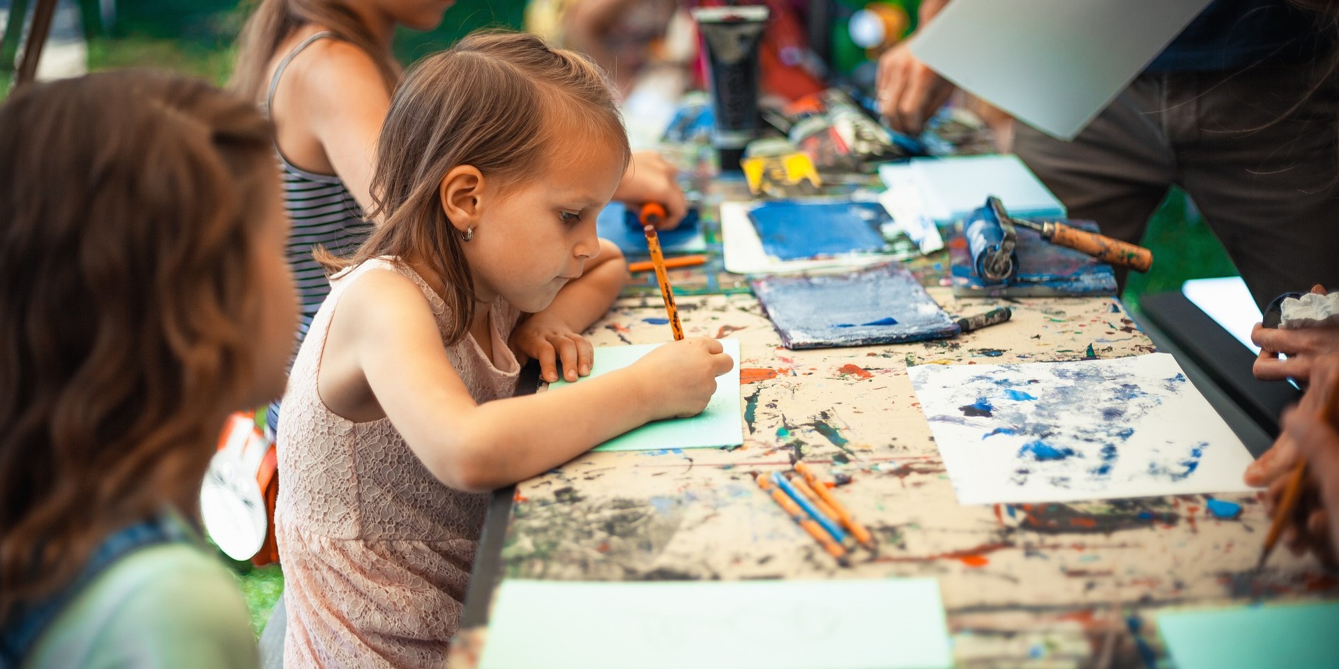 Ein kleines Mädchen sitzt an einem Tisch und malt mit Buntstiften. Es sind weitere Kinder im Hintergrund zu sehen.