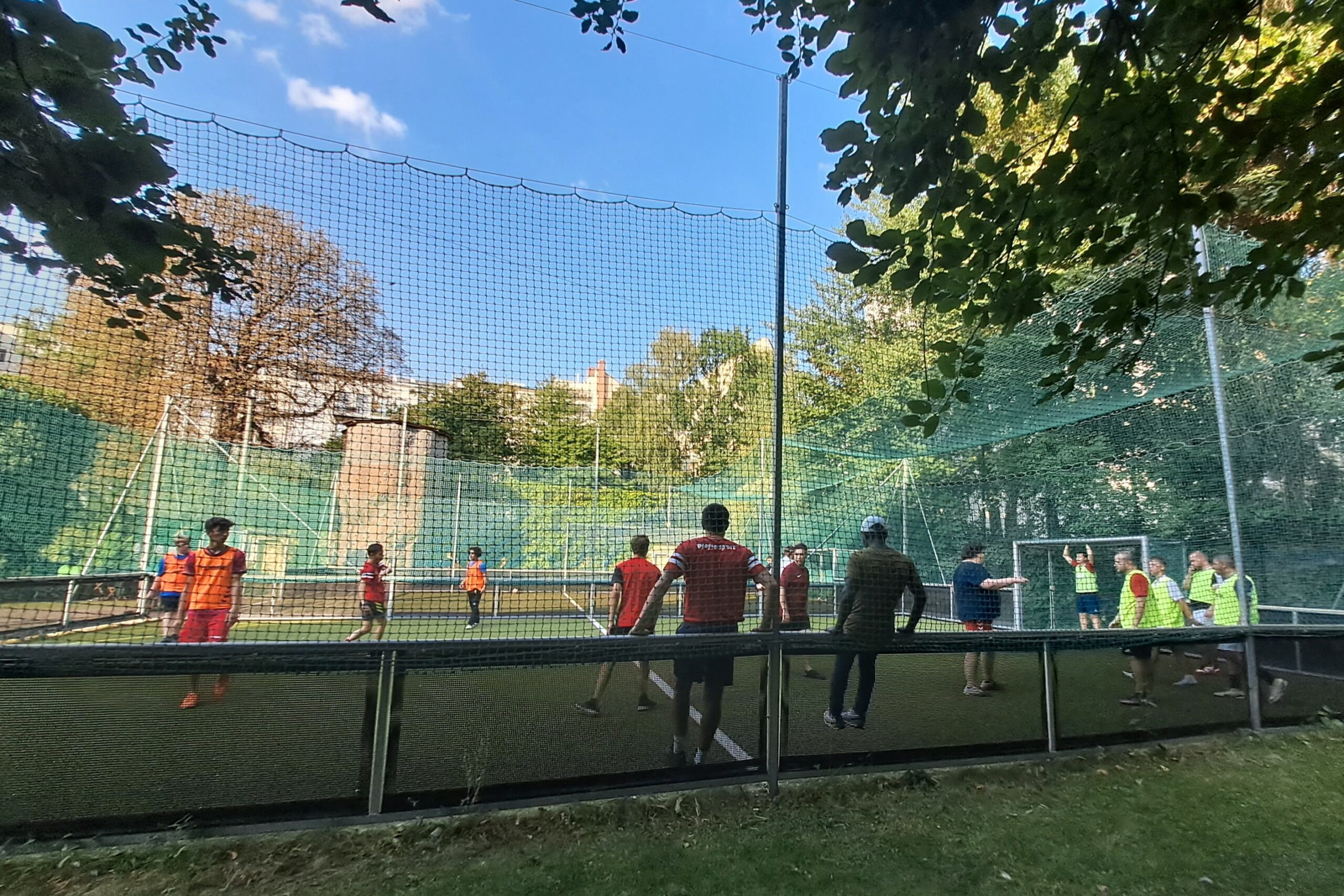 Ein kleines Fußballfeld mit Kunstrasen. Etwa ein Dutzend Spieler tragen gelbe und orangefarbene Westen und befinden sich auf dem Spielfeld. Am Spielfeldrand stehen zwei Trainer und beobachten das Spiel.