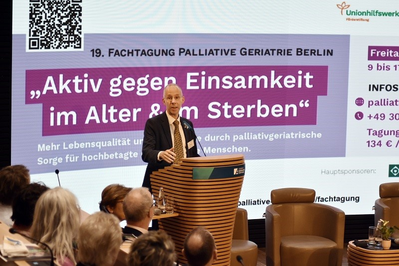 Ein Mann hält auf einer Konferenz eine Rede. Im Hintergrund ist eine Präsentation zu sehen: 19. Fachtagung Palliative Geriatrie Berlin "Aktiv gegen Einsamkeit im Alter & im Sterben"