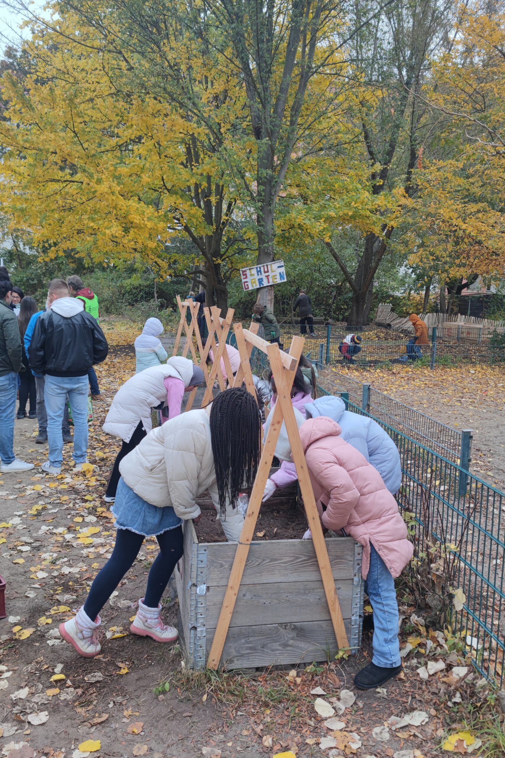 Rund acht Kinder gärtnern in einem Hochbeet auf einem Schulhof. Sie tragen dicke Winterjacken.