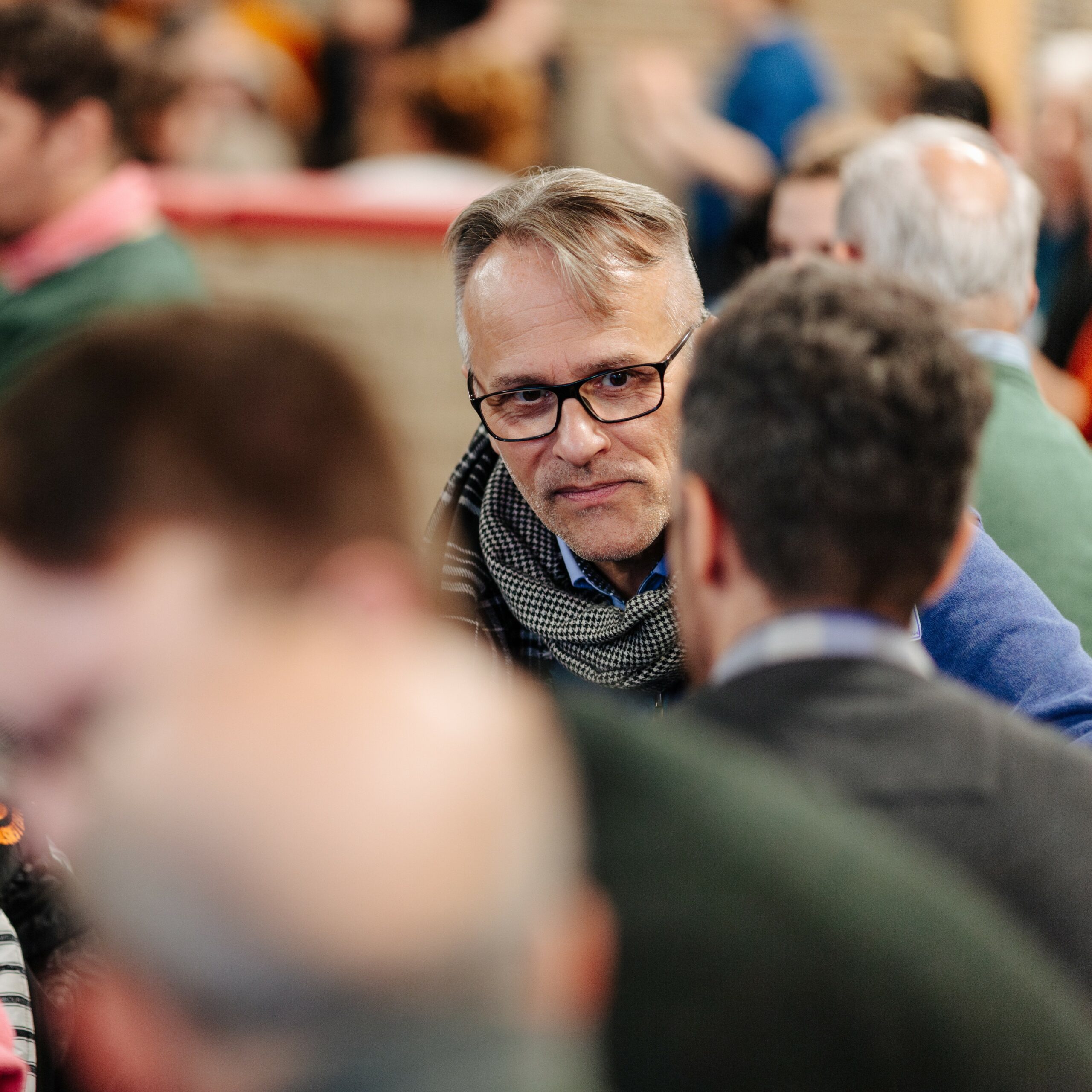 Bild einer Konferenz. Im Mittelpunkt ist ein Mann mit Brille zu sehen, der mit einem Mann spricht, der nur von hinten zu sehen ist. Weitere Personen sind in Gespräche vertieft.
