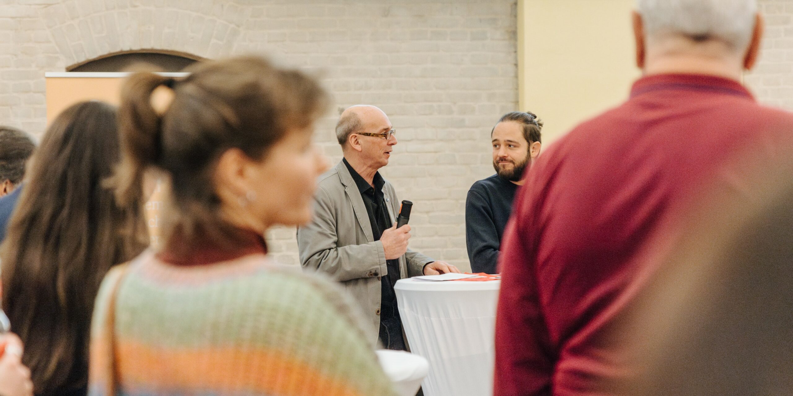 Bei einer Veranstaltung stehen zwei Männer an einem Stehtisch, einer von ihnen spricht ins Mikrofon. Im Vordergrund sind die Rücken mehrerer Zuschauer:innen zu sehen.