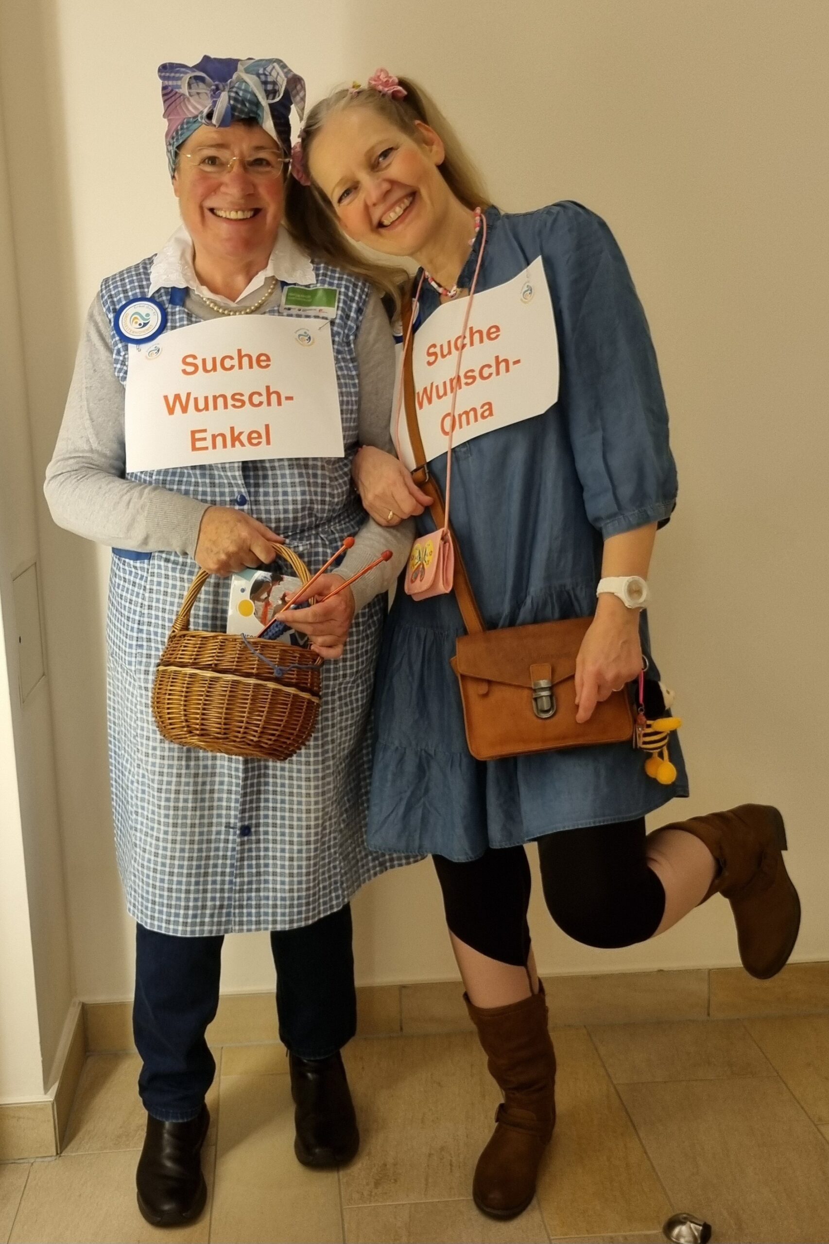 Helga und Stefanie vom Großelterndienst lachen in die Kamera. Sie sind verkleidet – Helga als Großmutter, Stefanie als Enkelin. Beide tragen ein Schild: Helga mit ‚Suche Wunsch-Enkel‘, Stefanie mit ‚Suche Wunsch-Oma‘.