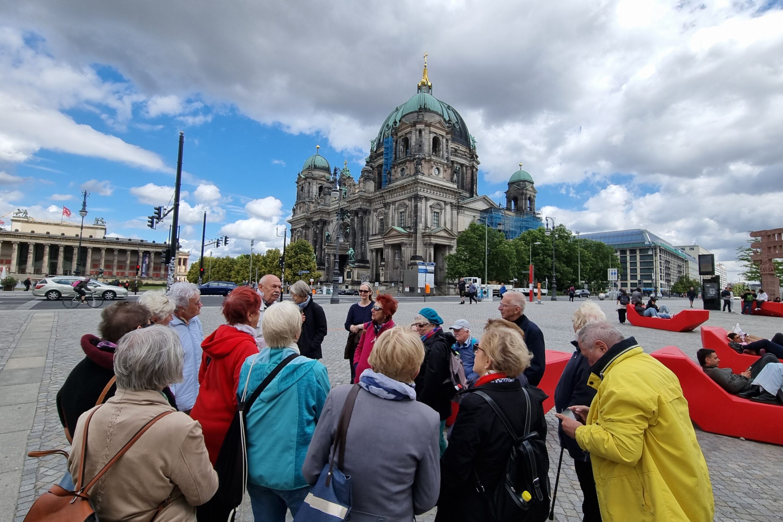 Eine Gruppe Senior:innen steht auf der Museumsinsel und hört einem Mann, vermutlich dem Tourguide, zu. Im Hintergrund ist der Berliner Dom zu sehen.