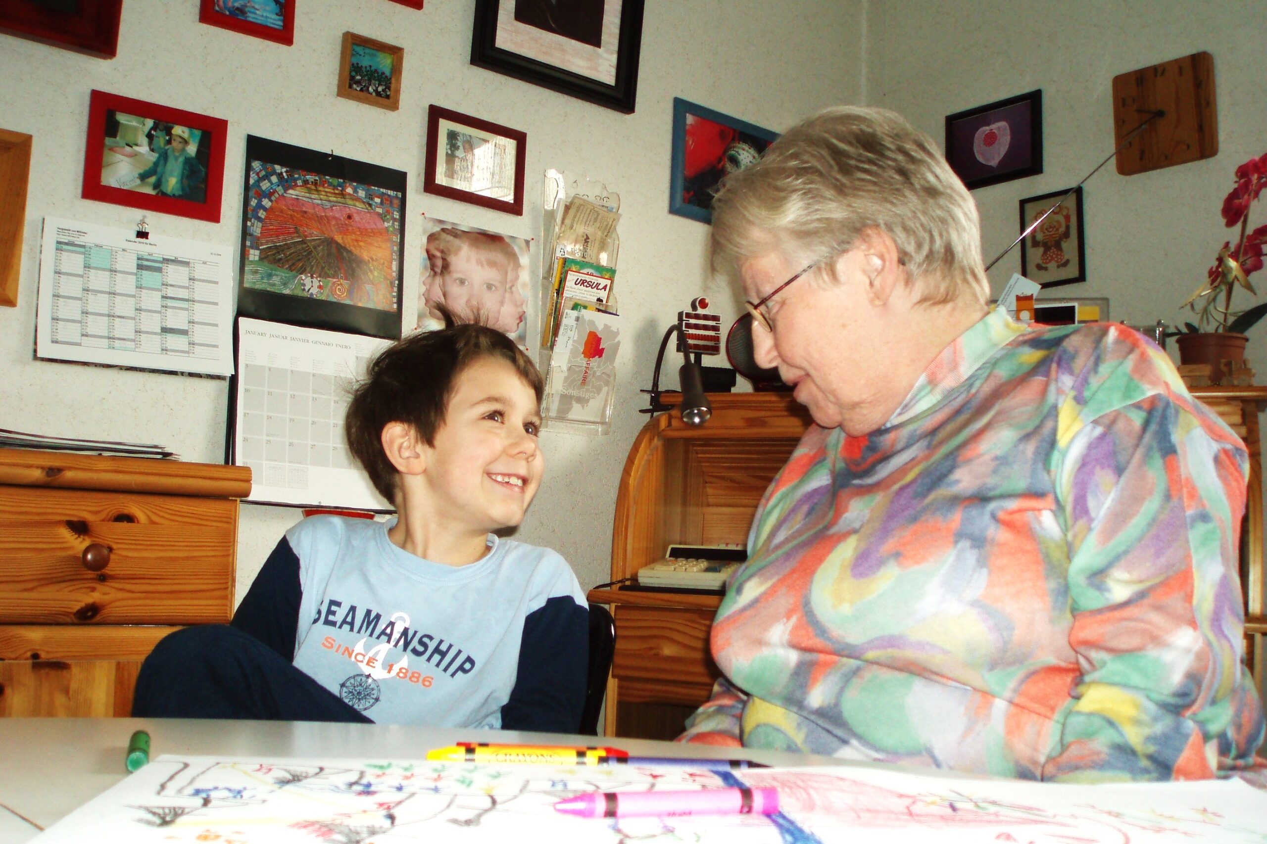 Eine ältere Frau sitzt zusammen mit einem Jungen in einem Zimmer, vermutlich im Wohnzimmer. Sie sprechen miteinander, während auf dem Tisch Malsachen liegen. Im Hintergrund sind viele Bilder an der Wand zu sehen.
