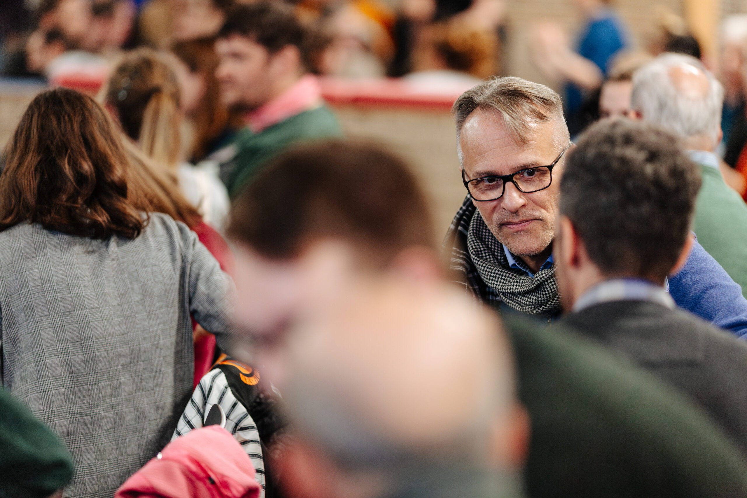 Bild einer Konferenz. Im Mittelpunkt ist ein Mann mit Brille zu sehen, der mit einem Mann spricht, der nur von hinten zu sehen ist. Weitere Personen sind in Gespräche vertieft.