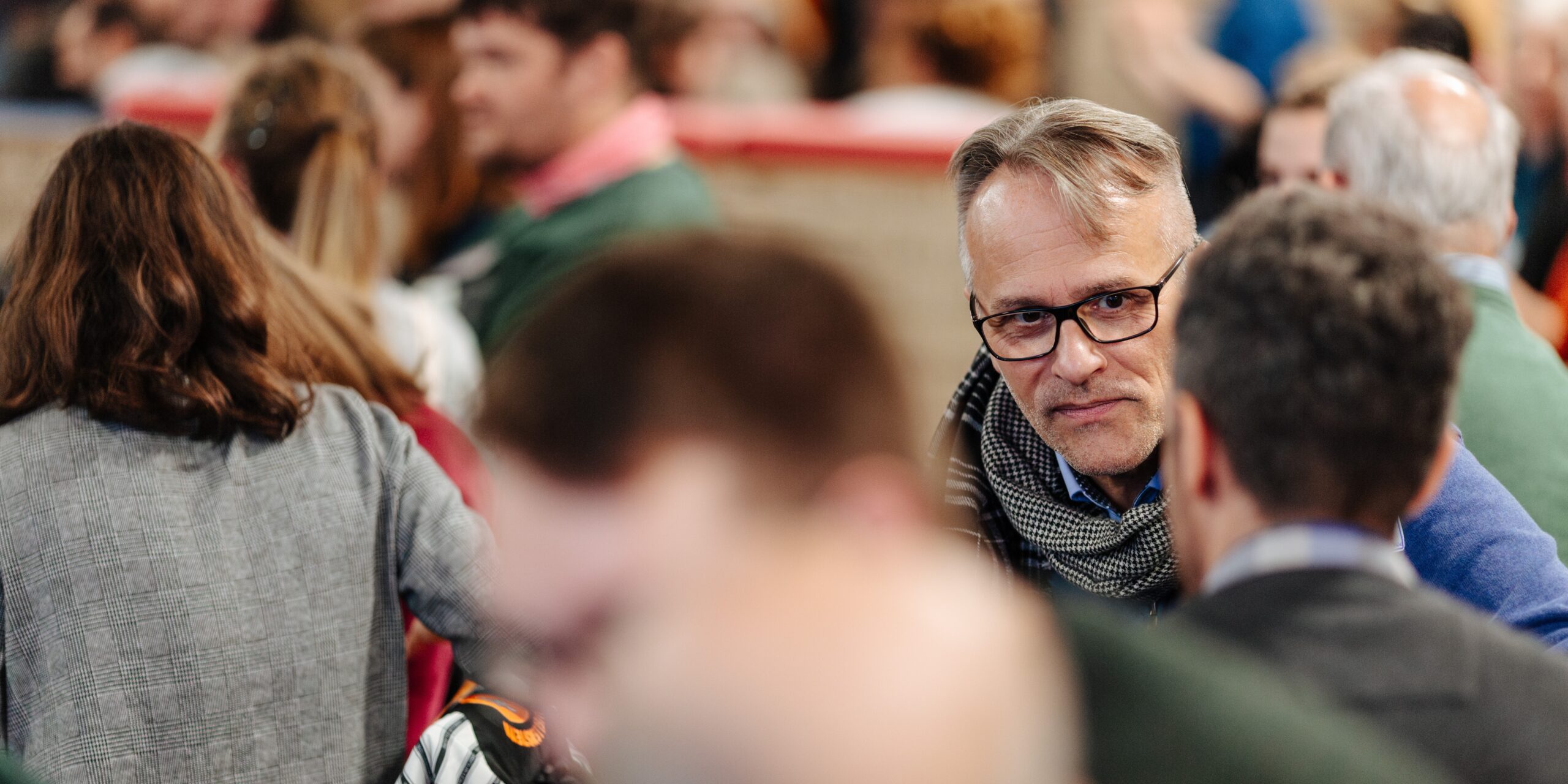 Bild einer Konferenz. Im Mittelpunkt ist ein Mann mit Brille zu sehen, der mit einem Mann spricht, der nur von hinten zu sehen ist. Weitere Personen sind in Gespräche vertieft.